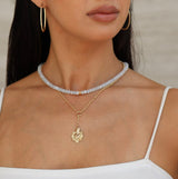 Woman wearing gold necklace with sacred heart and hoop earrings against a neutral background
